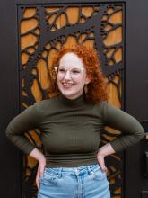 A red haired woman wearing a green turtleneck shirt, blue jeans, and glasses is smiling in front of a wood door with metal filigree. 