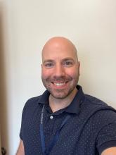 Picture of male, bald with short beard standing in front of white wall.